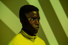 Young African American Male Athlete In Sportswear Looking Away While Standing Under Green Neon Illumination Near Corner With Striped Shadows In Studio