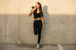© Luis Tenza/ADDICTIVE STOCK - Full body young slim female jogger in black sportswear with towel on neck leaning against concrete wall and drinking nutritive beverage after training on street
