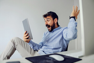 Young amazed bearded man dressed business casual sitting in office and looking at tablet.