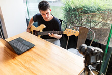 Focused Young Male Musician Sitting Near Window And Performing Music On Bass Guitar