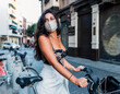© Jose Carlos Gutierrez/ADDICTIVE STOCK - Side view of charming young Latin female in stylish outfit and protective mask standing near bicycle on city street and looking away