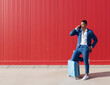 © Jose Carlos Gutierrez/ADDICTIVE STOCK - Full body of young African American man in elegant blue suit with luggage messaging on mobile phone while standing against red wall on street