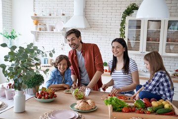 Enjoying weekend. Young beautiful family preparing a salad together in the modern kitchen at home. Mother and father teaching two little kids how to cut fresh vegetables