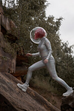 Low Angle Of Positive Young Female With Dyed Red Hair Wearing Silver Space Suit And Glass Helmet And Looking Away While Walking On Rocky Formation