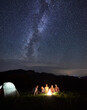 Night camping. Vertical shot group of tourists having a rest around campfire in the mountains in summer, having small talks near illuminated tent under beautiful sky full of stars and Milky way