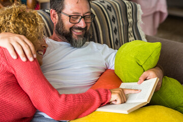 Adult people caucasian man and woman couple at home reading a book together sharing time and life on the couch - happy relationship lifestyle concept time for cheerful husband and wife