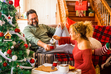 Wall Mural - Christmas time at home with happy adult caucasian couple having breakfast on the couch near the christmas tree - concept of family and love life together in traditional leisure activity indoor