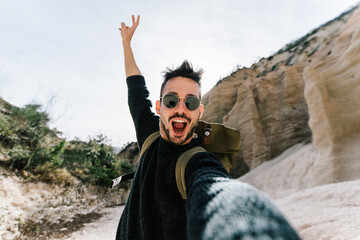 Wall Mural - Happy young man wearing backpack hiking on a mountain taking a selfie with smartphone and action cam.