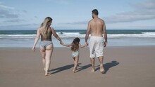 Parents And Kid Wearing Swimsuits, Walking On Beach To Water. Girl Holding Parents Hands, Jumping And Throwing Legs Up. Rear View. Family Outdoor Activities Concept