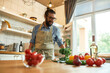 © Svitlana - Cheerful young man, Italian cook in apron holding basil leaves while getting ready to prepare healthy meal with vegetables, standing in the kitchen. Cooking at home concept