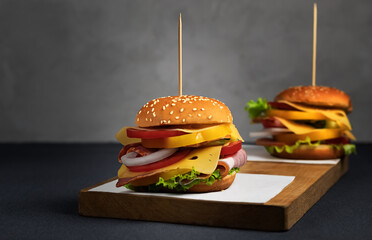 Burger with double cheese, bacon, lettuce, tomatoes and bell peppers on an old wooden board. Dark gray background. The second burger in blur. Side view. Copy space.