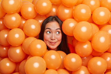 Holiday and entertainment concept. Curious cheerful pensive young woman with long dark hair sticks out head through orange inflated balloons enjoys free time organizes party or special event