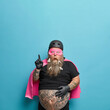 © wayhome.studio  - Emotional bearded European man in carnival superhero costume points above at blank space over blue wall wears clothespins on thick beard shows shocking advertisement keeps hand on fat tattooed stomach