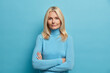© WHstudio Leushin N - Self confident serious calm middle aged experienced blonde woman stands with arms folded dressed in casual turtleneck shows professional vibe stands in assertive pose against blue background.