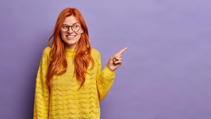 Wall Mural - Beautiful redhead young woman indicates finger at empty space with cheerful expression shows something nice dressed in casual yellow jumper isolated on purple background makes nice presentation
