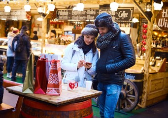 Canvas Print - Young couple doing christmas shopping at christmas fair, using mobilephone.