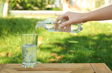 Wall Mural - Woman pouring water from bottle into glass at wooden table outdoors, closeup