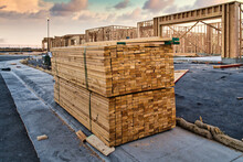Pallet Of 2x4 Studs For House Framing - Lumber Sitting On The Sidewalk Of A Residential Construction Site With Home Frames In The Background During Sunset With Beautiful Clouds