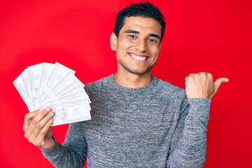 Wall Mural - Young handsome hispanic man holding money bag with dollar symbol pointing thumb up to the side smiling happy with open mouth