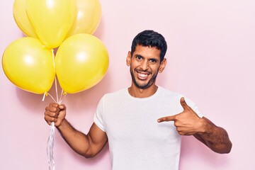 Young latin man holding balloons pointing finger to one self smiling happy and proud