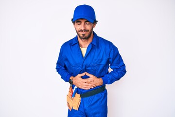 handsome young man with curly hair and bear weaing handyman uniform with hand on stomach because ind