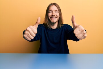 Wall Mural - Handsome caucasian man with long hair wearing casual clothes sitting on the table approving doing positive gesture with hand, thumbs up smiling and happy for success. winner gesture.