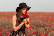 Beautiful Long Hair Woman In Black With Hat On Head At The Middle Of Field Full Of Red Flowers