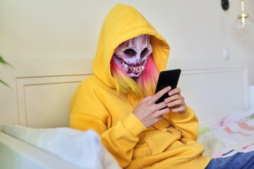 Halloween day, girl in scary face mask with hood makes video call on smartphone