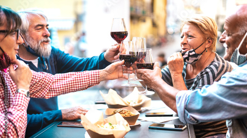 Senior couples toasting red wine at restaurant bar with face masks - New normal lifestyle concept with happy people having fun together at bar outdoors - Bright filter with focus on central glasses