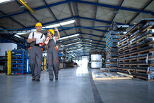 Factory Workers In Work Wear And Yellow Helmets Walking Through Industrial Production Hall And Discussing About Organization.