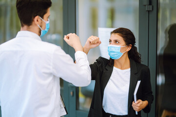 Portrait of two colleagues wearing face masks bumping elbows while greeting each other at work in office. Teamwork during pandemic in quarantine city. Covid-19.
