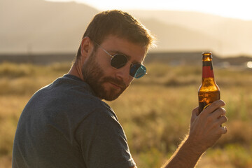 A young man, with glasses and a beard, drinks beer from a bottle, while enjoying the landscape and the sunset, in Galicia, northern Spain. Cheers!