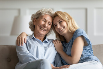 Tender closeness. Two females of different generatons adult mother and grown child cuddling on sofa, millennial woman former student embracing with care and affection happy grey-haired lady teacher