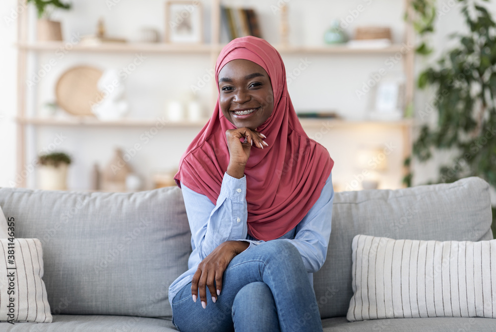 Joyful young african muslim woman in hijab posing on couch at home - obrazy, fototapety, plakaty 