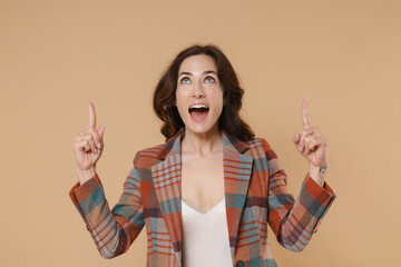 Wall Mural - Shocked surprised excited young brunette woman 20s wearing casual checkered jacket posing pointing index fingers up on mock up copy space isolated on pastel beige colour background, studio portrait.