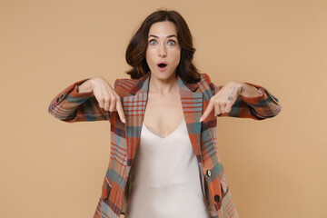 Wall Mural - Shocked surprised amazed young brunette woman 20s wearing casual checkered jacket posing pointing index fingers down on mock up copy space isolated on pastel beige colour background, studio portrait.