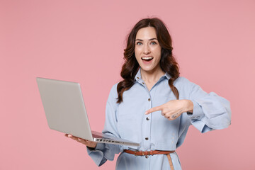 Excited young brunette woman 20s wearing casual blue shirt dress posing standing pointing index finger on laptop pc computer looking camera isolated on pastel pink colour background, studio portrait.