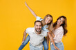 © ViDi Studio - Laughing young parents mom dad with child kid daughter teen girl in basic t-shirts giving piggyback ride to joyful, sitting on back isolated on yellow background studio portrait. Family day concept.