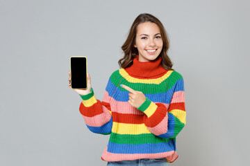 Wall Mural - Smiling young brunette woman in casual colorful knitted sweater point index finger on mobile cell phone with blank empty screen mock up copy space isolated on grey colour background, studio portrait.