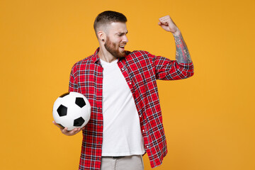 Shocked strong young man football fan in basic red shirt cheer up support favorite team with soccer ball showing biceps muscles isolated on yellow background studio. People sport leisure concept.