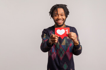 Extremely happy african male blogger holding in hand social media heart sign pointing at you, like button, follow us. Indoor studio shot isolated on gray background