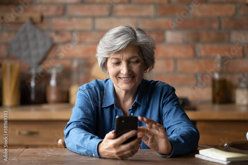 Fotografía Smiling modern elderly 60s woman sit at table at home kitchen text or message on smartphone gadget