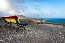 An Empty Bench, Painted In A Rainbow Of Colors And A View Of Playa De Sotavento De Jandia. Fuerteventura. Canary Islands. Spain.