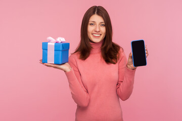 Happy brunette woman in pink sweater holding and showing mobile phone and blue gift box. Indoor studio shot isolated on pink background