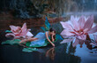 A beautiful woman a little cute fairy with butterfly wings lies on green water lily leaf. Fantasy scenery of huge pink flowers on the lake. River nymph, girl pixie in an blue dress girl touching water