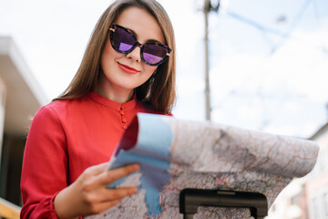 Shot of a beautiful young woman traveler in sunglasses, keeping map in hands and looking for the sightseeing sitting on the bus stop. Concept of traveling, vacation, using map.