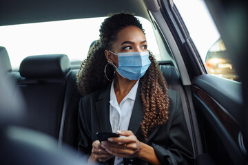 Young business woman in a mask checking her mobile cell phone on a backseat of a taxi during covid-19 quarantine. Business trips during pandemic, new normal and coronavirus travel safety concept.