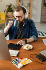 Wall Mural - Young creative male freelance designer in headphones having coffee with cookies