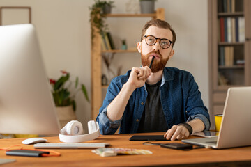 Wall Mural - Young bearded pensive freelancer with stylus by his mouth working at home