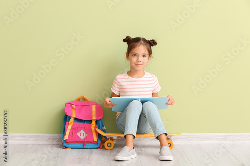 Fotografía Little schoolgirl with skateboard and notebook sitting near color wall